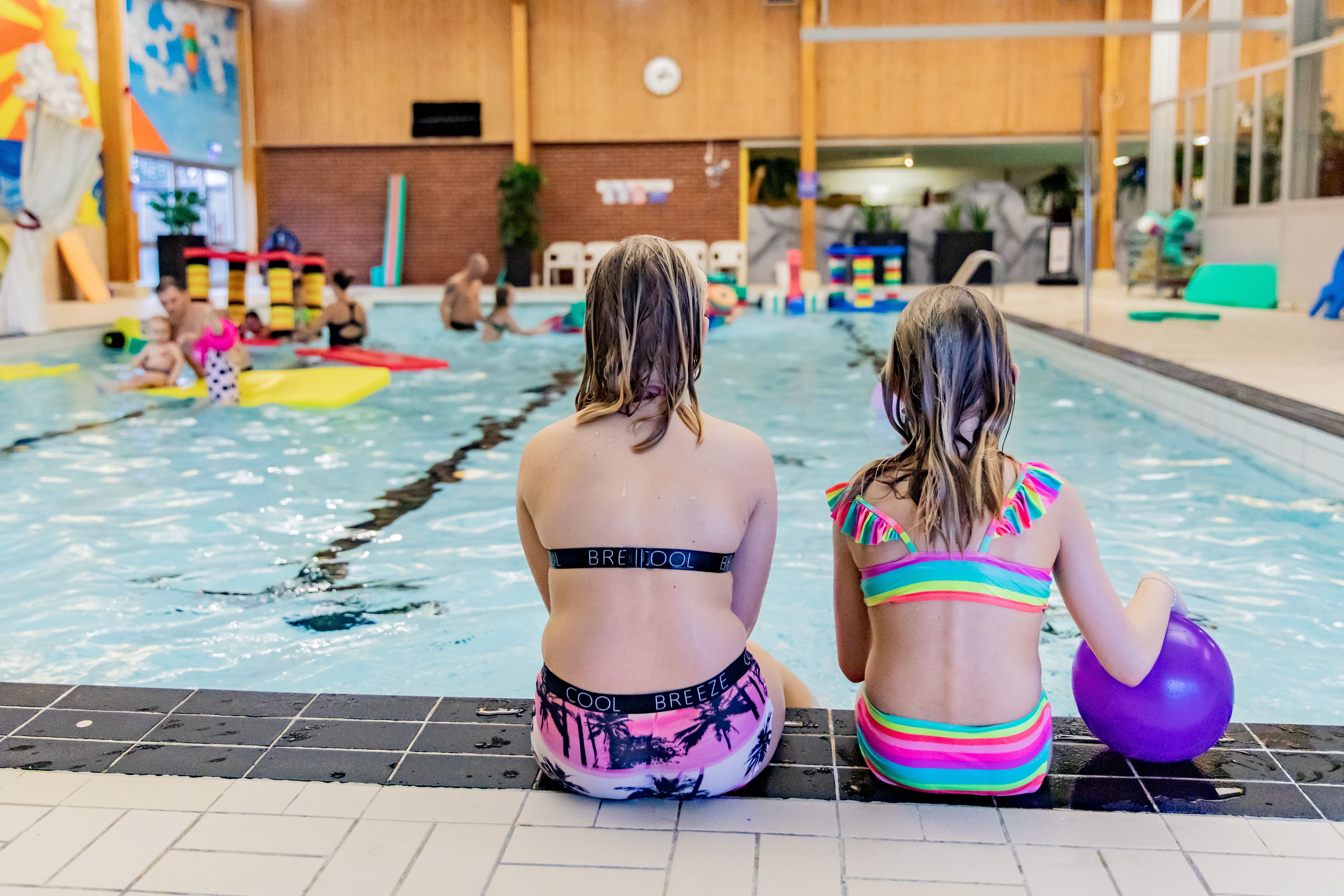 Två flickor med färgglada badkläder sitter på bassängkanten med ryggen mot kameran. den en flickan håller i en lila boll. I bassängen är det lekande människor och flytleksaker.